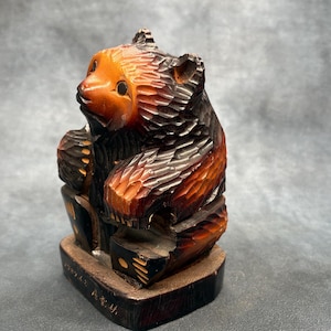 Peut inclure: Figurine d'ours en bois sculpté à la main, avec une surface texturée détaillée. L'ours est représenté en position assise, avec un corps brun rougeâtre et une tête de couleur orange clair. La base est brun foncé, et le style de sculpture suggère un art populaire.