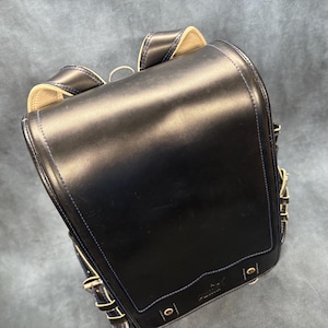 May include: A black leather backpack with a rectangular shape and a flap closure. The backpack has gold-colored hardware, white stitching, and a small logo on the front. The straps are adjustable.