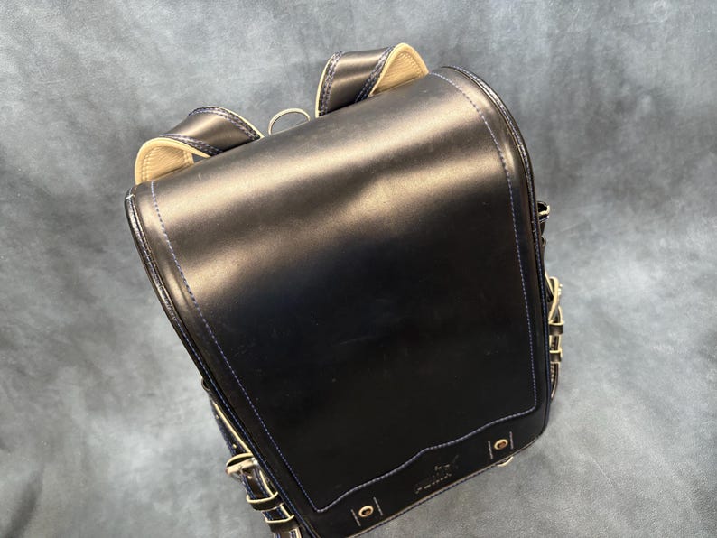 May include: A black leather backpack with a rectangular shape and a flap closure. The backpack has gold-colored hardware, white stitching, and a small logo on the front. The straps are adjustable.