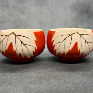 May include: Two ceramic bowls with a white interior and a vibrant orange exterior. Each bowl features a leaf design in black and white. The bowls are small and round, with a footed base.
