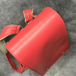 May include: A red leather school backpack with adjustable straps and a flap closure. The backpack has a classic design and is made of high-quality leather.