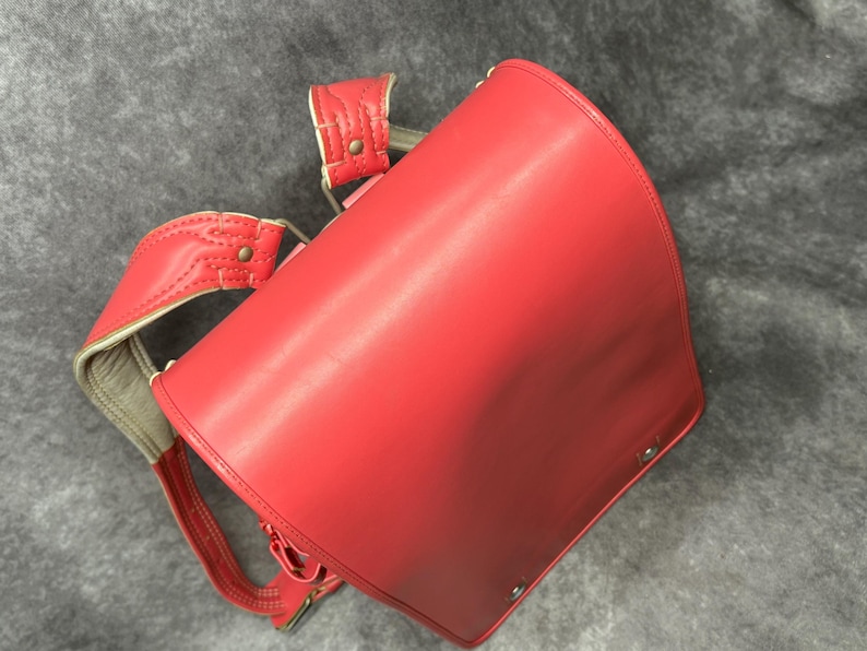 May include: A red leather school backpack with adjustable straps and a flap closure. The backpack has a classic design and is made of high-quality leather.