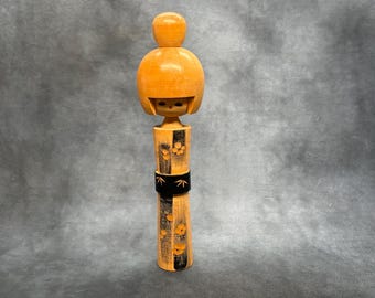 May include: A wooden Kokeshi doll featuring a smooth, light-colored rounded head with a topknot and a cylindrical body. The body is decorated with vertical black stripes, orange floral patterns, and a central black band with white bamboo leaf motifs.