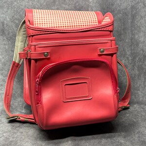 May include: A red leather backpack with a white and red gingham pattern on the top flap. The backpack has two large compartments and a smaller pocket on the front. The backpack has adjustable straps and a handle on the top.
