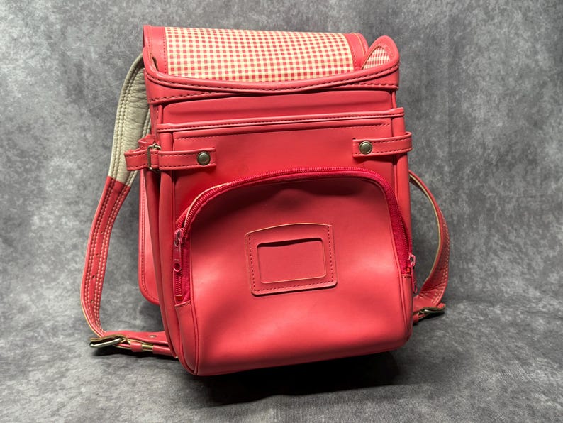 May include: A red leather backpack with a white and red gingham pattern on the top flap. The backpack has two large compartments and a smaller pocket on the front. The backpack has adjustable straps and a handle on the top.