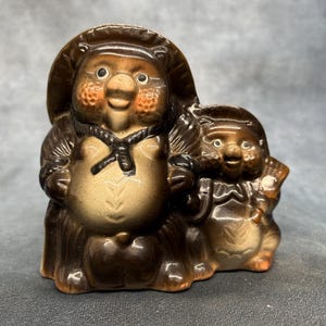 Peut inclure: Figurine en céramique de Tanuki, un chien viverrin japonais traditionnel, dans des tons bruns et beiges. La plus grande figurine porte un chapeau et a une expression joviale, avec une plus petite figurine à côté. La figurine a une finition brillante.
