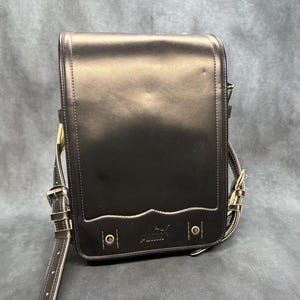 May include: Black leather satchel with a flap closure and adjustable shoulder strap. The bag features a decorative wave design along the bottom edge and a Puma logo. The bag has metal hardware and a buckle closure.