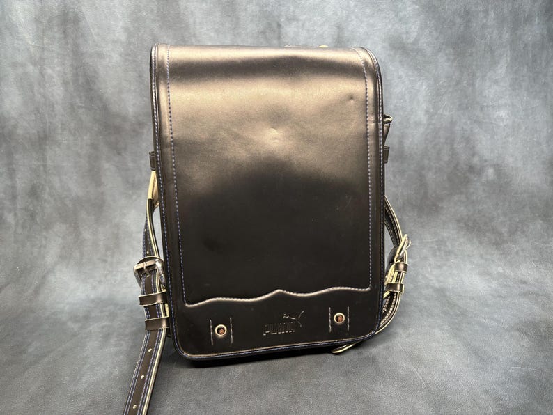 May include: Black leather satchel with a flap closure and adjustable shoulder strap. The bag features a decorative wave design along the bottom edge and a Puma logo. The bag has metal hardware and a buckle closure.