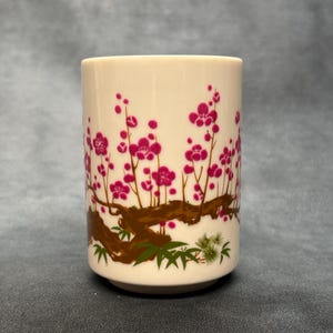 May include: A cylindrical off-white ceramic cup featuring a decorative wrap-around illustration of a brown tree branch with vibrant magenta plum blossoms and small green leaves at the base, presented in a simple, handleless, upright design.
