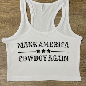 May include: White racerback tank top with the text "MAKE AMERICA COWBOY AGAIN" in black. The text is centered and features three stars between two horizontal lines. The tank top has a scoop neck and a racerback design.