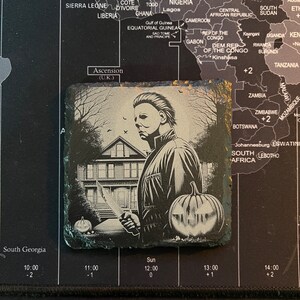 May include: A black and white image of a house with a spooky pumpkin and a man holding a knife. The man is wearing a black jacket and has a white mask on his face. The image is on a black slate coaster.