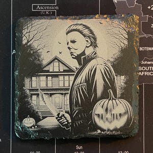 May include: Black and white illustration of a man wearing a jacket and holding a knife. He is standing in front of a house with a jack-o-lantern in the foreground. The image is on a black slate coaster.