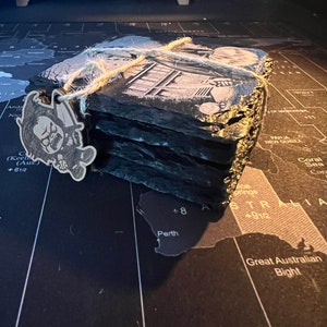May include: A stack of four black slate coasters with a printed image of a building. The coasters are tied together with twine and a small wooden tag with a cartoon image is attached.