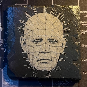 May include: A black slate coaster with a white image of a man's face with a grid of pins over it. The man has a serious expression and is looking directly at the viewer.