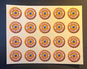 Puede incluir: Veinticinco pegatinas redondas con un diseño dorado y rojo. Las pegatinas presentan una estrella azul y el texto "Newcastle Brown Ale".