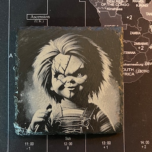 May include: A black and white image of a creepy doll with a knife. The doll is wearing a striped shirt and overalls. The background is dark and blurry.