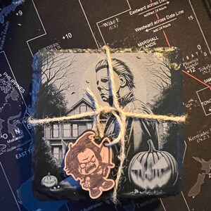 May include: Set of three black slate coasters featuring a horror movie theme. The coasters feature a black and white illustration of a house, a pumpkin, and a character from a horror movie. The coasters are tied together with twine and a small wooden charm of a character from a horror movie is attached.