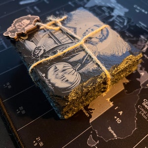 May include: A set of four black slate coasters with a spooky Halloween design featuring a person holding a pumpkin. The coasters are tied together with twine.