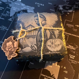 May include: A set of four black slate coasters featuring a black and white illustration of a horror movie character holding a knife and a pumpkin. The coasters are tied together with twine.