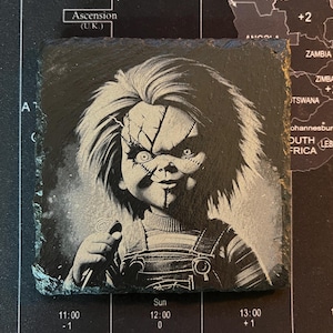 May include: Black and white image of a creepy doll with a knife. The doll has a stitched-up face and a mischievous grin. The image is printed on a black slate coaster.