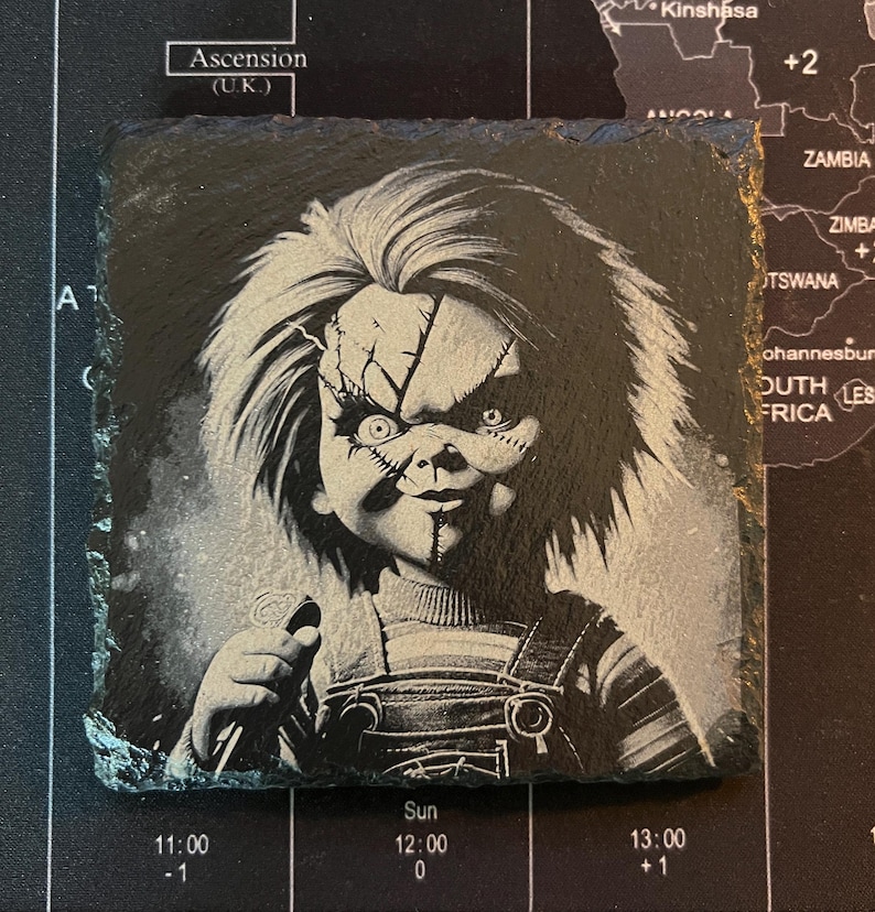 May include: Black and white image of a creepy doll with a knife. The doll has a stitched-up face and a mischievous grin. The image is printed on a black slate coaster.