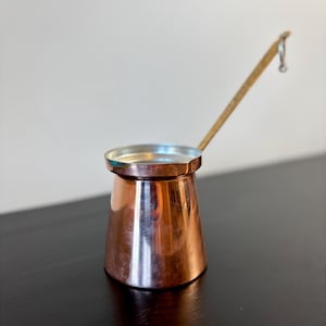May include: A copper coffee pot with a long brass handle. The pot has a rounded base and a flared top. The handle has a small loop at the end.