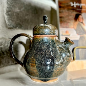 Peut inclure: Une théière en céramique de couleur vert foncé et marron avec un corps arrondi et un petit bec verseur. La théière a un glaçage moucheté et une surface légèrement irrégulière.