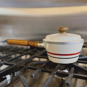 May include: White enamel saucepan with a wooden handle and a red and black stripe design. The saucepan is sitting on a black stovetop.