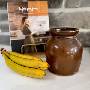 May include: A brown ceramic jar with a rounded body and a slightly flared rim. Beside it are three ripe bananas and a cookbook with the title "3 fois par jour". The items are arranged on a white countertop.