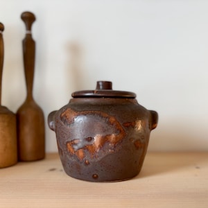 Peut inclure: Un pot en céramique brun avec un couvercle. Le pot a un glaçage brun marbré avec une teinte légèrement orange. Le pot a deux petites poignées sur les côtés.
