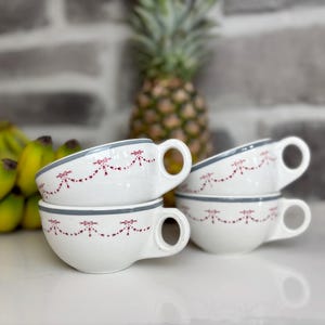 May include: Four white ceramic teacups with gray rims and handles are stacked. Each cup is decorated with a repeating pattern of red bows. A pineapple and bananas are in the background.