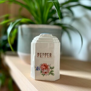 Peut inclure: Saliere en céramique blanche avec un motif floral et l'inscription "PEPPER" en lettres dorées.