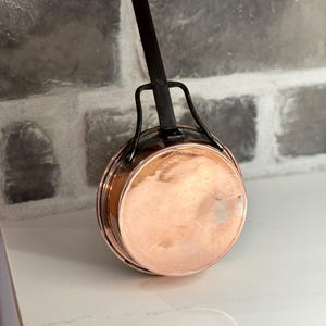 Peut inclure: Une casserole en cuivre avec une poignée en fer noir. La casserole est ronde et a une surface lisse et brillante.