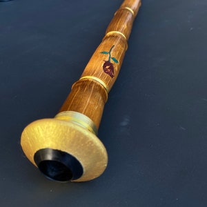 May include: A long, wooden musical instrument with a bamboo-like design. The instrument features a rose design with green leaves and gold accents. The end has a gold-colored flared base with a black center.