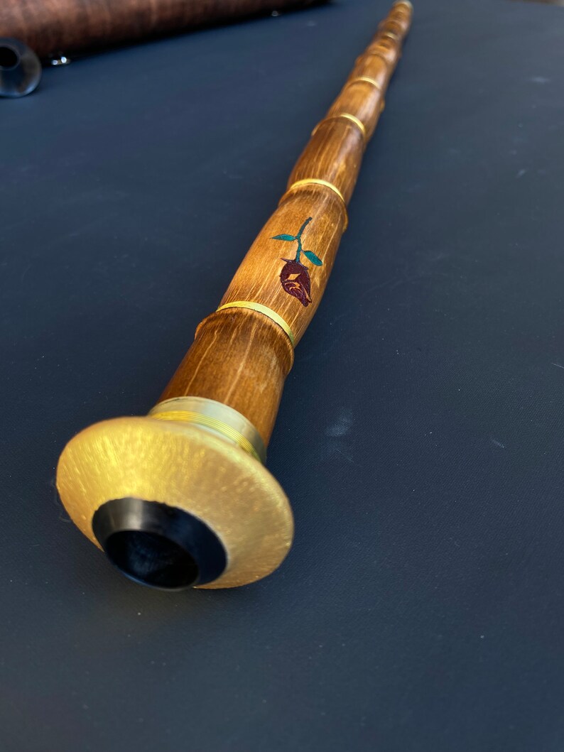 May include: A long, wooden musical instrument with a bamboo-like design. The instrument features a rose design with green leaves and gold accents. The end has a gold-colored flared base with a black center.