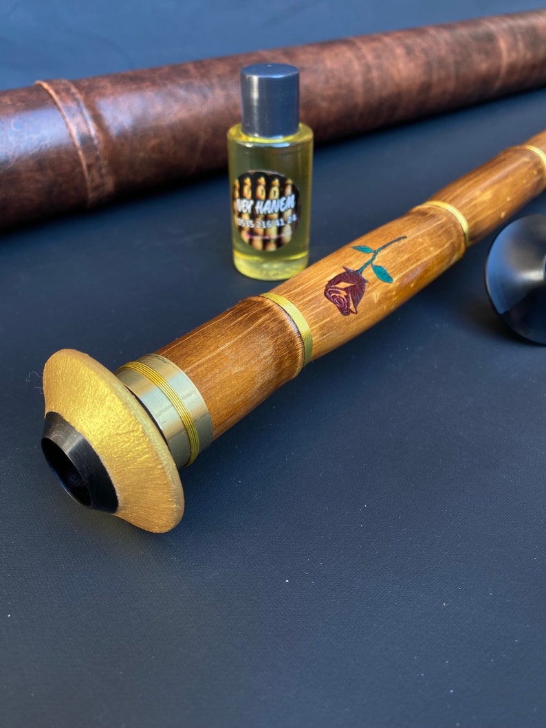 May include: A wooden wind instrument with a gold-colored bell and a rose design. A small bottle of liquid with a label and a brown leather case are also present. The instrument has brass accents and a black mouthpiece.