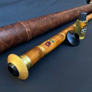 May include: A wooden wind instrument with a rose design, brass accents, and a black mouthpiece. A brown leather case and a small bottle of oil are also present. The instrument is likely a ney, a traditional end-blown flute.