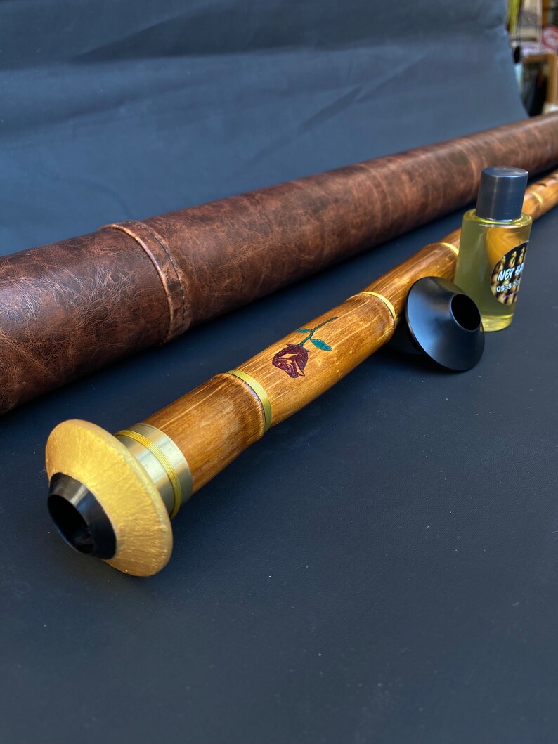 May include: A wooden wind instrument with a rose design, brass accents, and a black mouthpiece. A brown leather case and a small bottle of oil are also present. The instrument is likely a ney, a traditional end-blown flute.