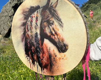 Puede incluir: Tambor redondo con una pintura de acuarela de la cabeza de un caballo adornada con plumas. El tambor tiene una superficie de piel natural y está adornado con tela gris. Cintas rosas cuelgan de la parte inferior. El fondo muestra una escena soleada al aire libre.