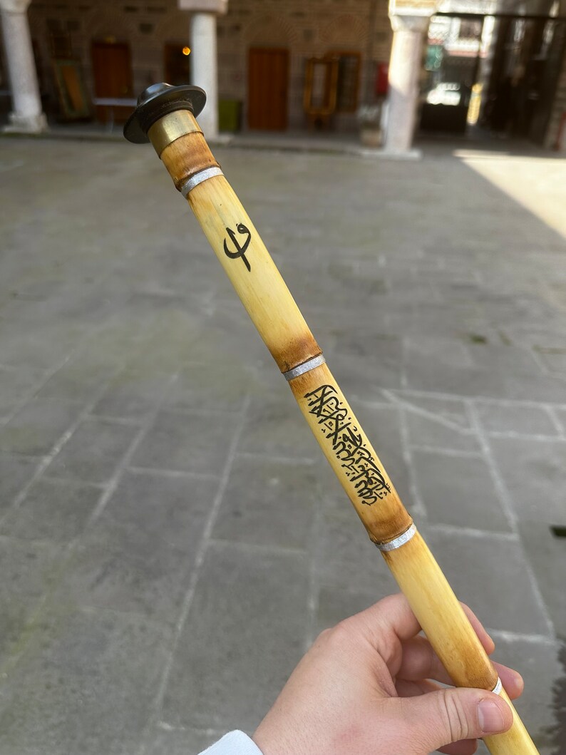 Może przedstawiać: Tradycyjny flet trzcinowy, czyli ney, z metalową nasadką i ozdobnymi opaskami. Bambusowy instrument posiada wytrawioną kaligrafię i symbol. Ney to instrument dęty używany w muzyce bliskowschodniej.