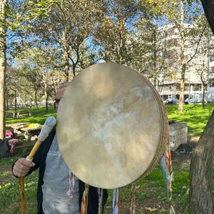 May include: A large, round drum with a light tan drumhead is held outdoors. The drum has a woven side and is decorated with colorful ribbons. A drumstick with a blue and white handle is held in front of the drum. The background shows a park with trees and green grass.