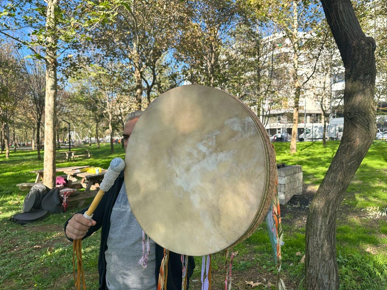 May include: A large, round drum with a light tan drumhead is held outdoors. The drum has a woven side and is decorated with colorful ribbons. A drumstick with a blue and white handle is held in front of the drum. The background shows a park with trees and green grass.