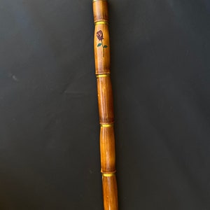 May include: A wooden walking cane with a rose design. The cane is made of bamboo-like segments, connected by brass-colored rings. The top has a rounded, light-colored wooden cap. A painted rose with green leaves is visible on the cane's shaft.