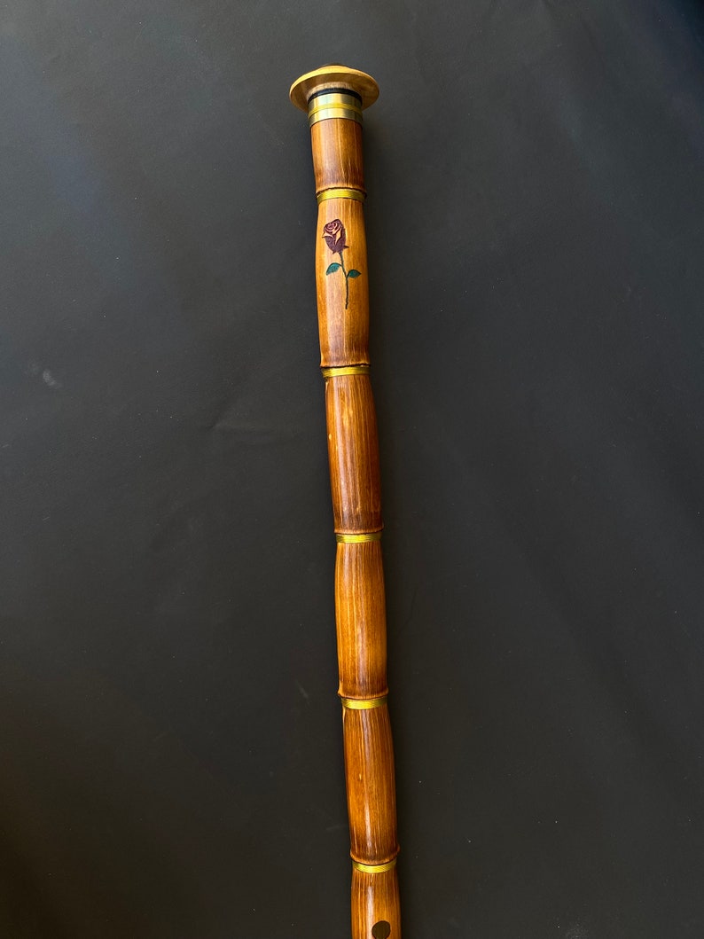 May include: A wooden walking cane with a rose design. The cane is made of bamboo-like segments, connected by brass-colored rings. The top has a rounded, light-colored wooden cap. A painted rose with green leaves is visible on the cane's shaft.