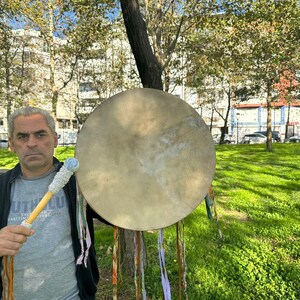 May include: A large, round, tan drum with a wooden handle and a white and blue drumstick. The drum is held in front of a person standing in a park with green grass and trees.