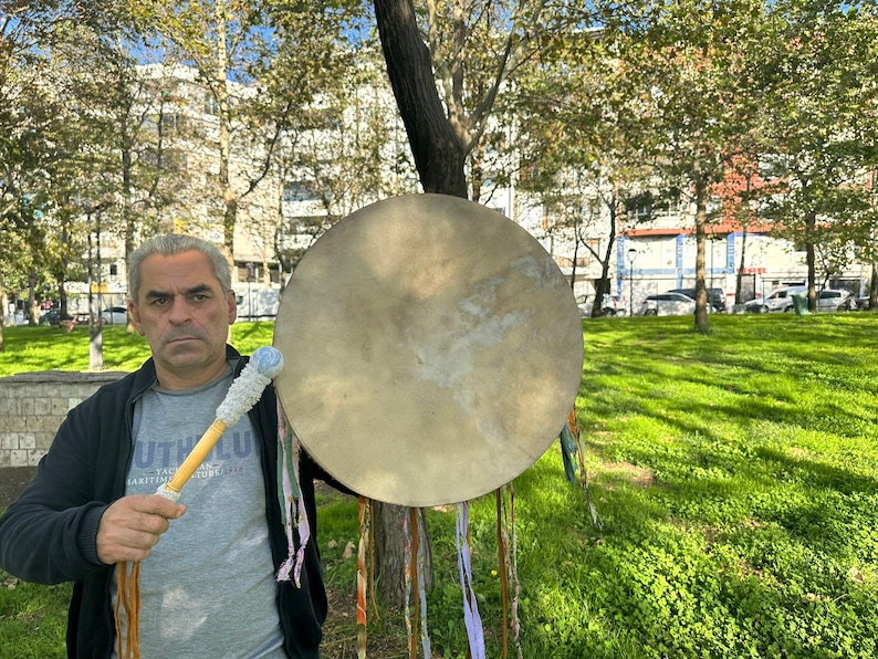 May include: A large, round, tan drum with a wooden handle and a white and blue drumstick. The drum is held in front of a person standing in a park with green grass and trees.