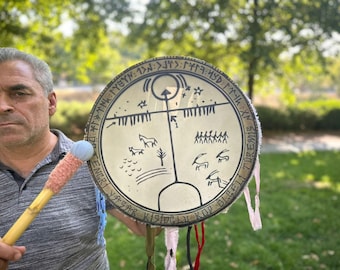 May include: A circular, beige drum with a central design of lines, symbols, and figures, held by a person. The drum has text around the edge and is accompanied by a mallet with a blue head and pink handle.