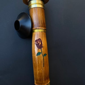 May include: A bamboo smoking pipe with a dark rose design. The pipe has a wooden mouthpiece and a black bowl. Brass rings accent the bamboo shaft. The pipe is a natural brown color.