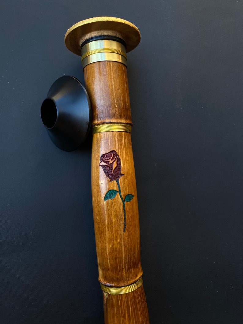 May include: A bamboo smoking pipe with a dark rose design. The pipe has a wooden mouthpiece and a black bowl. Brass rings accent the bamboo shaft. The pipe is a natural brown color.