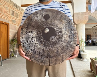 May include: A large, round, wooden art piece with a dark brown and gray color scheme. The artwork features a central design with a yin-yang symbol and a circular hole. The wood grain and cracks are visible, adding texture.